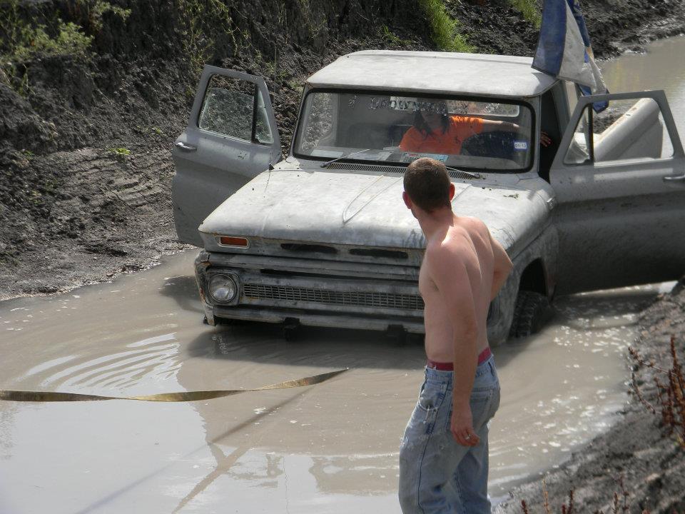 Guy watching a beat-up pickup truck stuck in a muddy ditch. One door is open