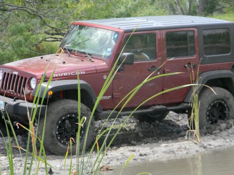 1989 Ford Bronco - A maroon Jeep Wrangler Rubicon navigates through a muddy, watery terrain with tall grass obscuring the view.