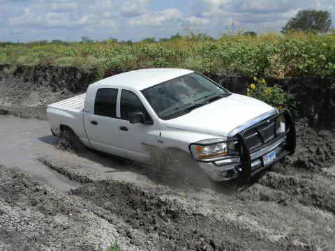 1989 Ford Bronco - A white pickup truck navigates a muddy off-road trail, kicking up dirt and water.