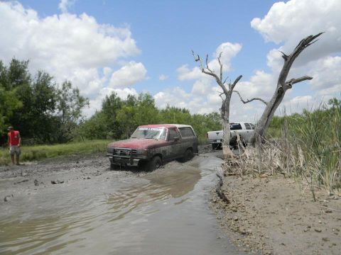1989 Ford Bronco - Two SUVs navigate a muddy path under a cloudy sky, with a person standing nearby.