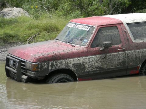 1989 Ford Bronco - This red and white SUV is partially submerged in muddy water, with mud splattered all over its body.