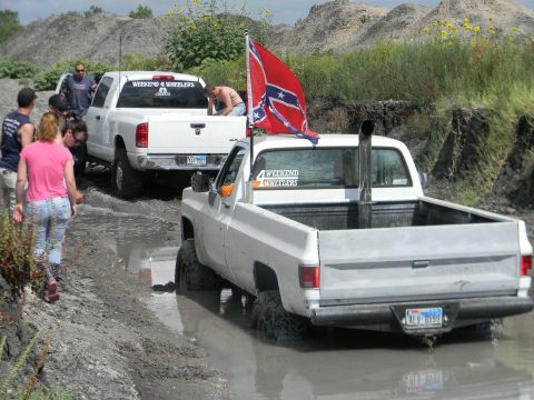 1989 Ford Bronco - Two trucks navigate a muddy off-road course, one flying a Confederate flag and sporting a prominent exhaust stack.