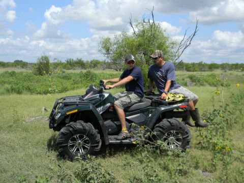 1989 Ford Bronco - Two men ride a green all-terrain vehicle through a grassy field with scattered trees and yellow flowers.