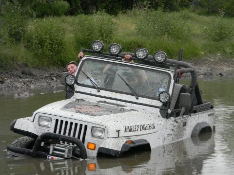 1989 Ford Bronco - A white Jeep Wrangler with "HARLEY DAVIDSON" text is stuck in muddy water with people inside.