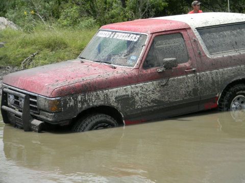 1989 Ford Bronco - A heavily muddied 1987 Ford Bronco is stuck in murky water, with a person visible in the background.