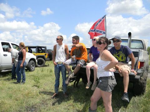 1989 Ford Bronco - A group of people gather around off-road vehicles under a cloudy sky, with a confederate flag flying.
