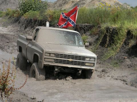 1989 Ford Bronco - Mud-splattered pickup truck navigates a muddy terrain, flying a Confederate flag.