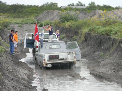 1989 Ford Bronco - Two pickup trucks navigate a muddy track, with people standing by and flags displayed.