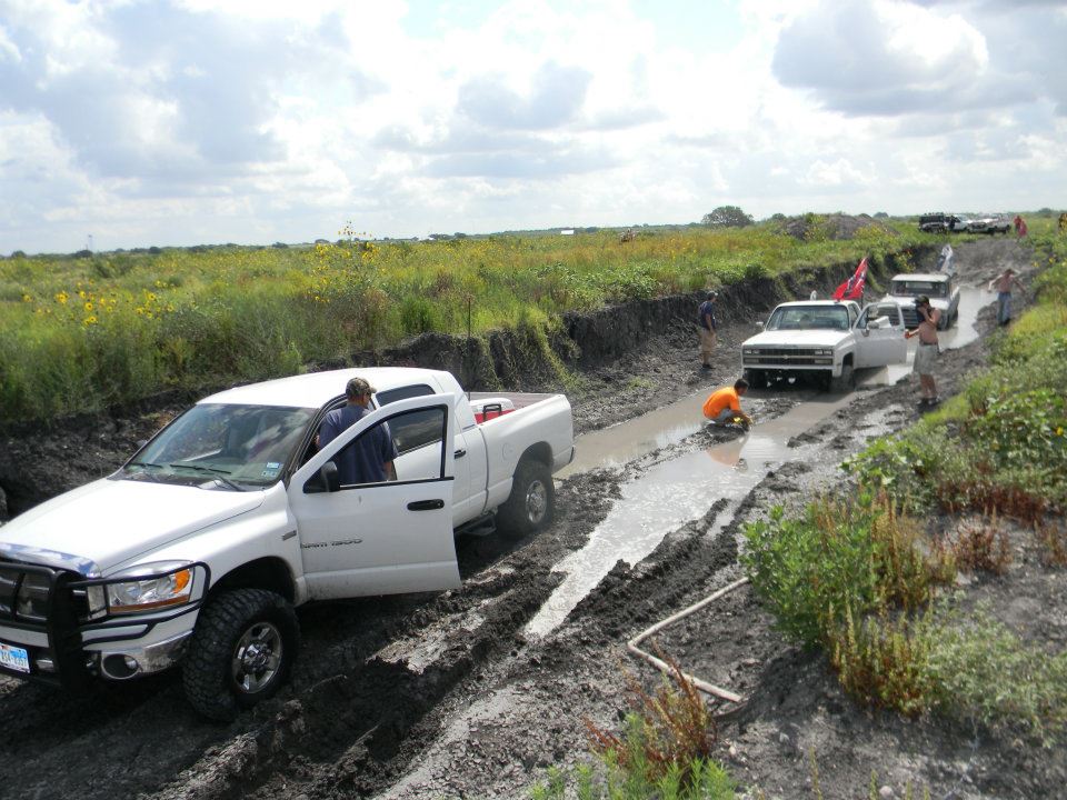 White pickup trucks driving through a muddy ditch with people around