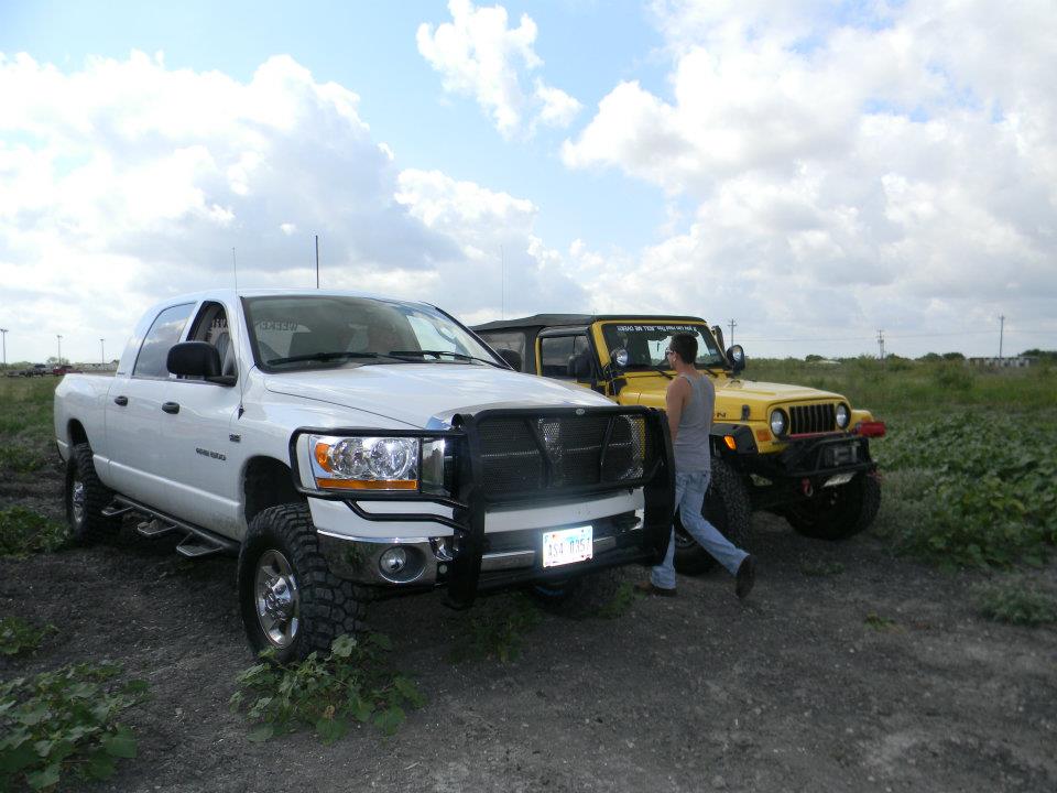 White pickup truck with black bull bar and yellow jeep parked on dirt with green vegetation under a cloudy sky