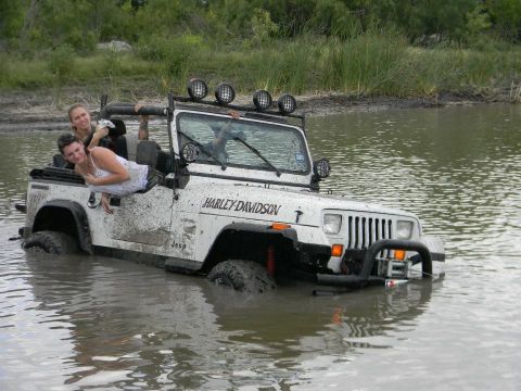 1989 Ford Bronco - A white Jeep Wrangler, adorned with a Harley Davidson decal, is partially submerged in muddy water with two women inside.