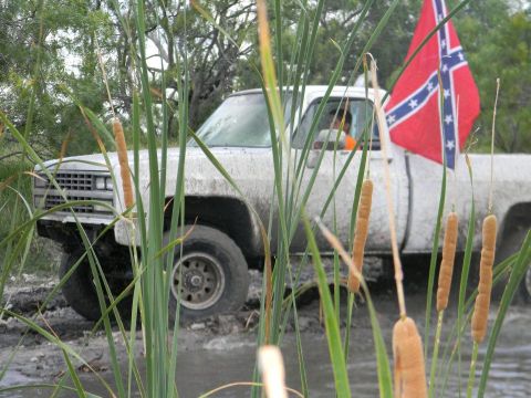 1989 Ford Bronco - A white pickup truck, possibly a 1989 model based on its grille, navigates a muddy terrain while flying a confederate flag.