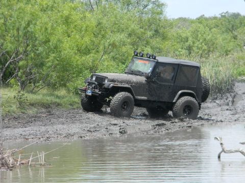 1989 Ford Bronco - A black Jeep Wrangler navigates through a muddy, watery terrain with lush green foliage in the background.