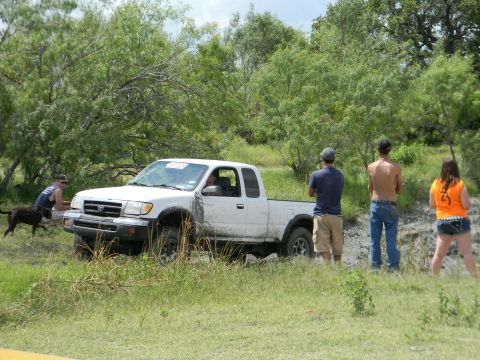 1989 Ford Bronco - A white pickup truck is stuck in the mud with onlookers observing the scene.