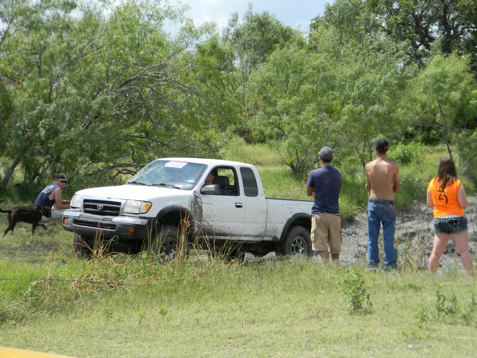 White pickup truck stuck in mud, front quarter view. Mud splattered on the side. People and a dog stand nearby