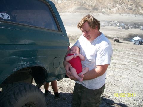 1991 Ford Bronco - Man holds a baby next to a green SUV with a dented rear quarter panel, with other vehicles in the background.