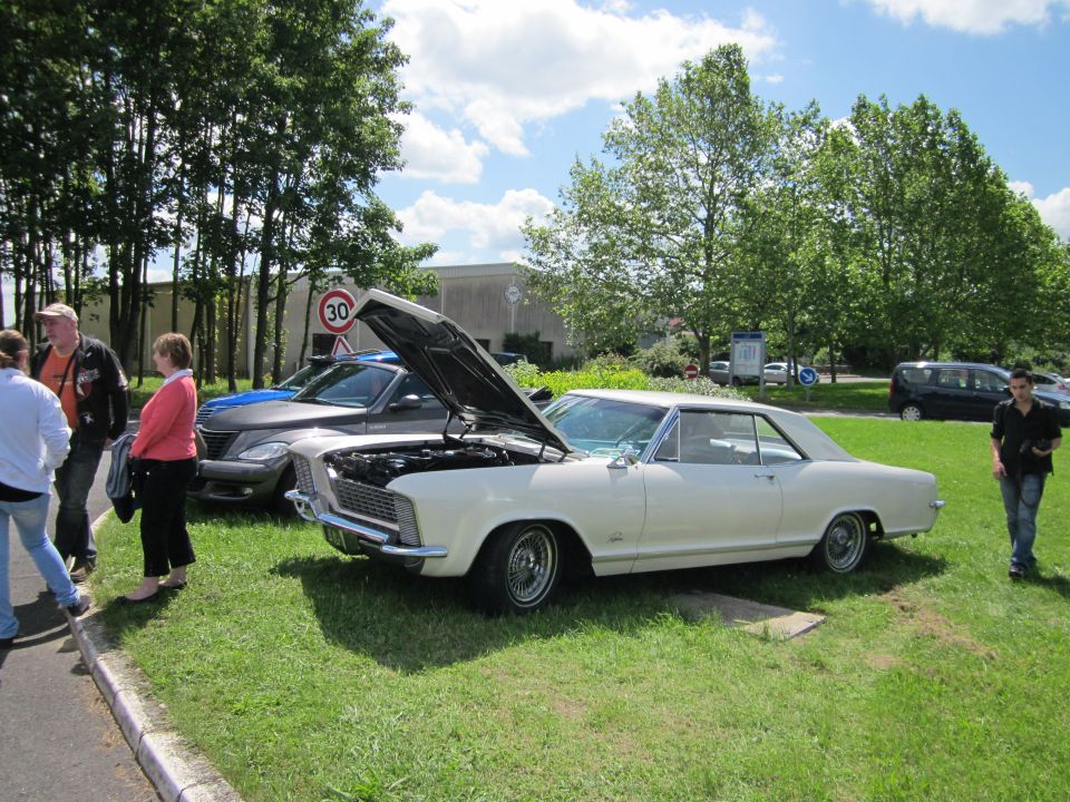 White 1965 Buick Riviera coupe with hood open, parked on grass next to a grey car. People stand nearby