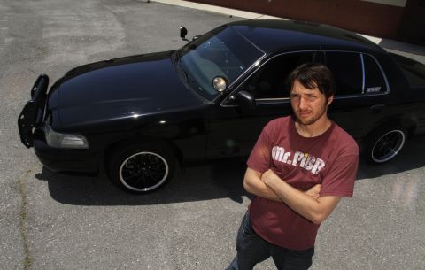 2003 Ford Crown Victoria - man stands arms crossed beside a black Ford sedan with black wheels and an open driver door