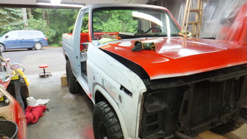 Ford Bronco body in primer and red paint, hood open, in a garage. Front grille removed