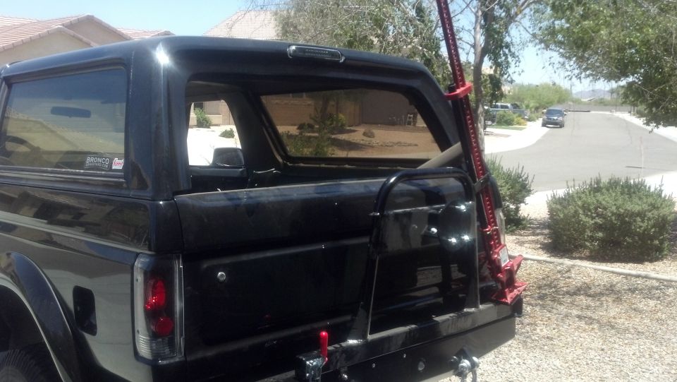 Rear of black SUV with spare tire carrier and red Hi-Lift jack mounted on the side. A sticker on the rear window says 'BRONCO'
