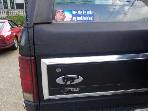 1986 Ford Bronco - A black Ford Bronco is seen from the rear, with a humorous sticker on the window and a red car in the background.