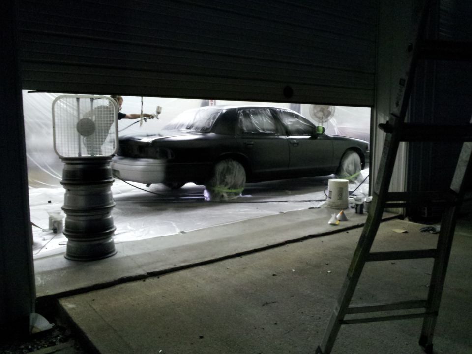 Car covered in plastic and tape, being spray painted inside a garage with a fan and ladder nearby