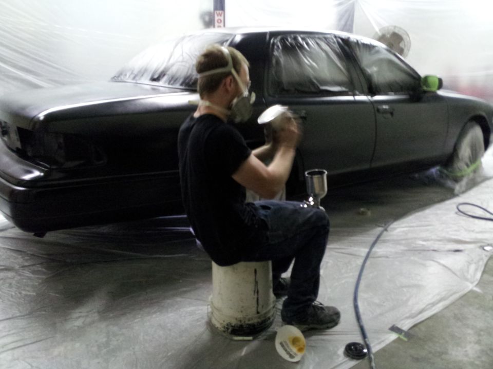 Man in mask sprays black paint onto a car covered in plastic sheeting
