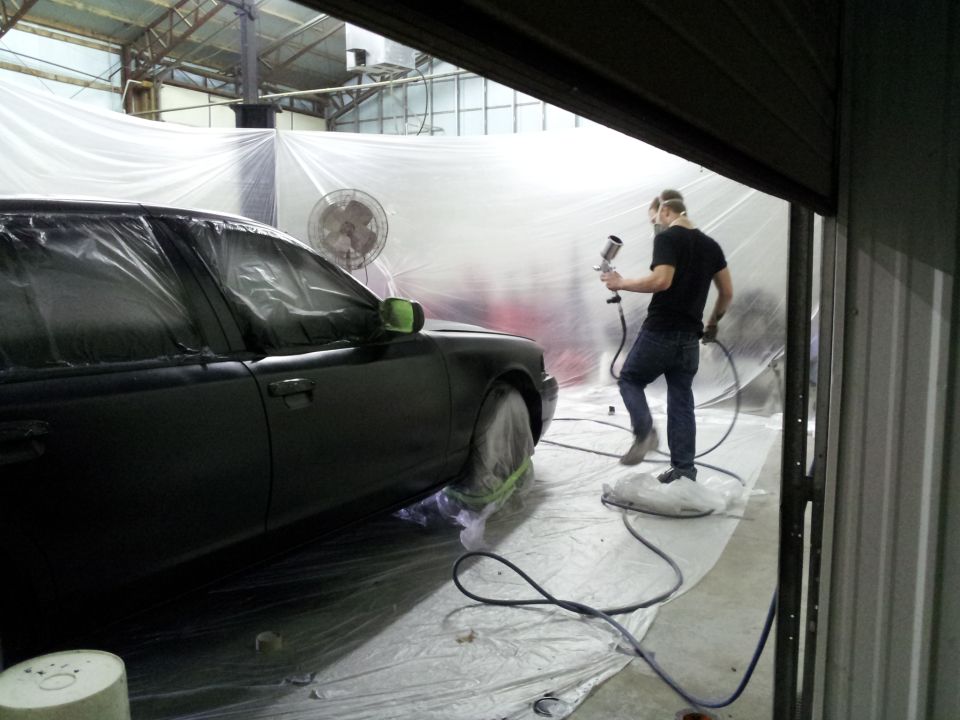 Black car covered in plastic, being prepped for paint in a spray booth with a man holding a spray gun