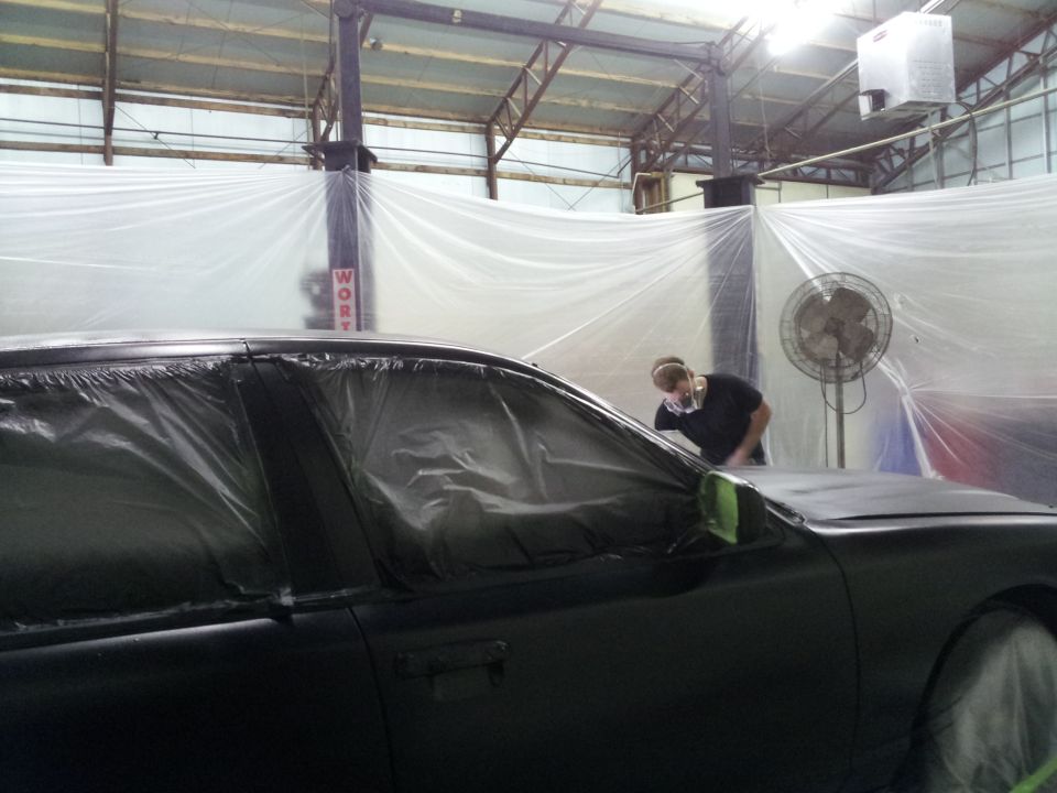 Car covered in plastic sheeting in a paint booth with a person wearing a mask working on it