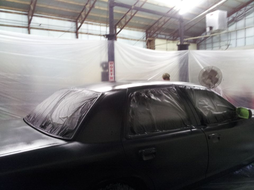 Black sedan covered in plastic sheeting in a paint booth, side view