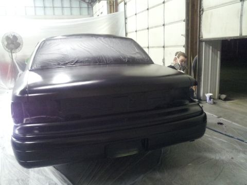 1994 Mercury Grand Marquis - A person in a mask works on painting a black sedan, which is covered in protective plastic.