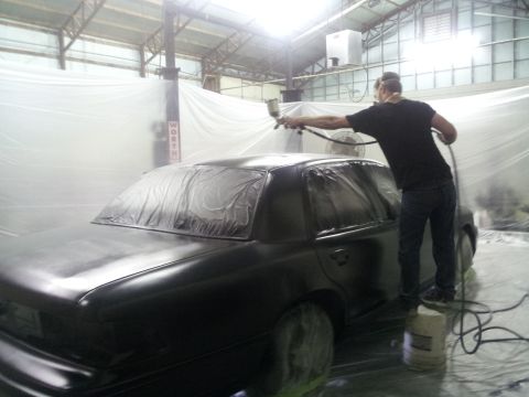 1994 Mercury Grand Marquis - A person sprays paint onto a black sedan in a workshop, with plastic sheeting covering the surroundings.
