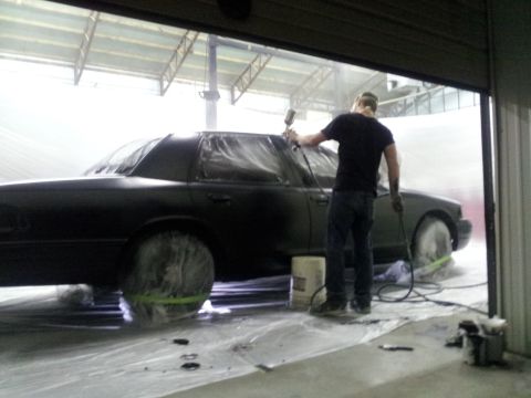 1994 Mercury Grand Marquis - A person sprays paint onto a black sedan that is covered in plastic sheeting inside a spray booth.