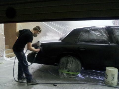 1994 Mercury Grand Marquis - A person sprays paint onto a black sedan covered in plastic sheeting.