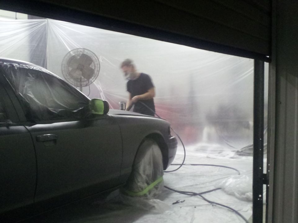 Dark sedan covered in plastic sheeting in a paint booth with a fan and a person