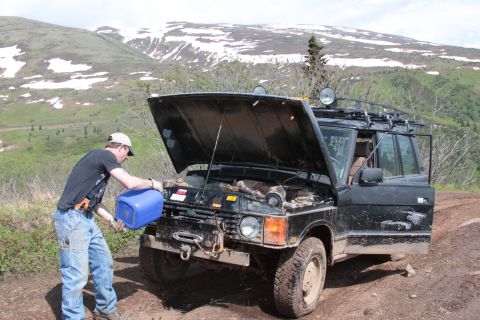 2005 Jeep TJ - A person tends to the engine of a vehicle parked on a dirt road with a mountainous landscape behind it.