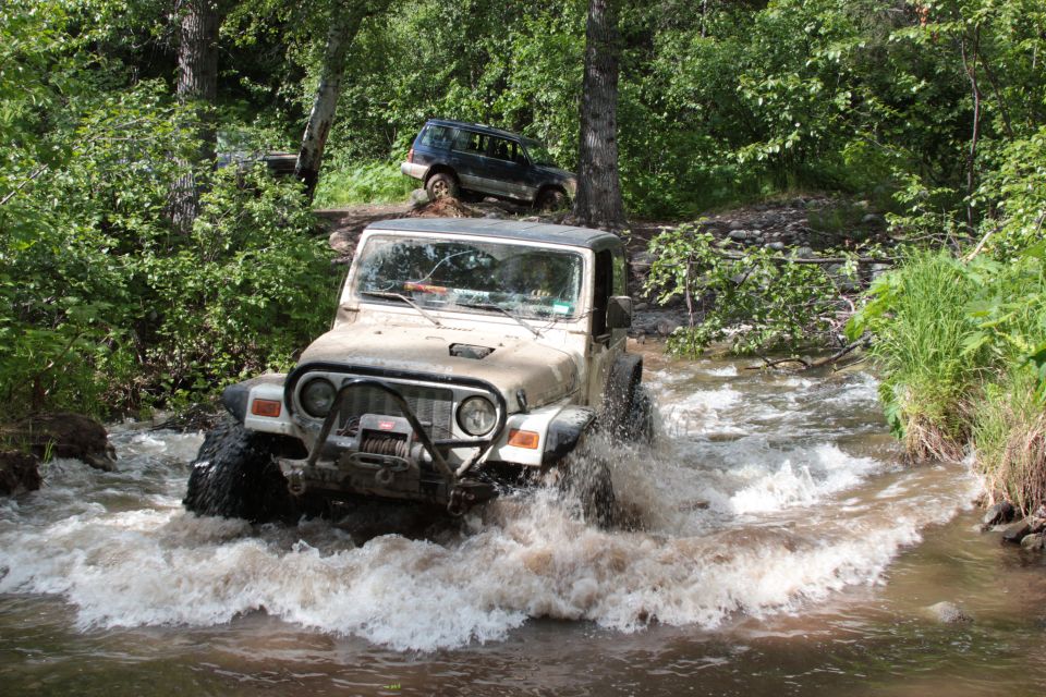 2005 Jeep TJ driving through a shallow stream with another SUV on the bank behind it
