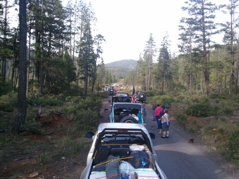 1994 Ford Bronco - A group of off-road vehicles travels down a wooded dirt road, carrying camping gear and displaying American flags.