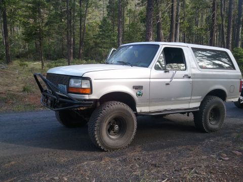 1994 Ford Bronco - 1994 Ford Bronco with aftermarket bumper and tires sits on a dirt road surrounded by trees.