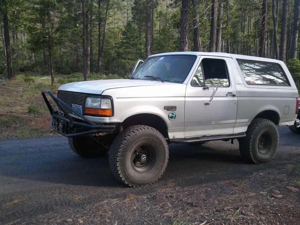 White 1994 Ford Bronco with aftermarket front bumper and off-road tires parked on a dirt road