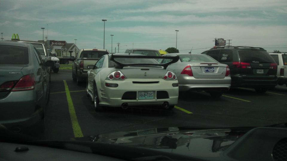 White Mitsubishi Eclipse with a large rear spoiler and custom body kit, parked in a lot. License plate HH4-TOS