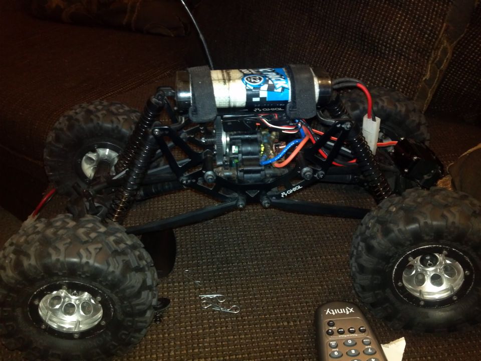 RC rock crawler with large tires, exposed motor, and battery pack. Remote control is visible in the foreground