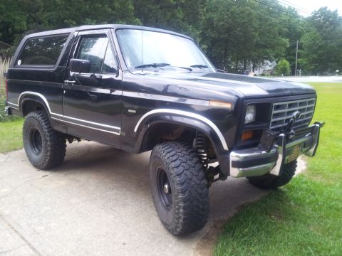1986 Ford Bronco - This lifted black Ford Bronco sits on a concrete driveway with grass and trees in the background.