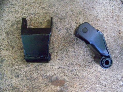 1985 Ford Bronco - Two black metal suspension parts are shown on a concrete surface. One part is a bracket, the other has a bushing.