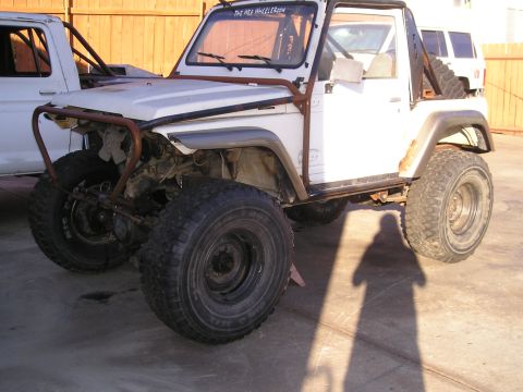 1987 Suzuki Samurai - This modified off-road vehicle features large tires and a prominent roll cage, with its front bumper removed.