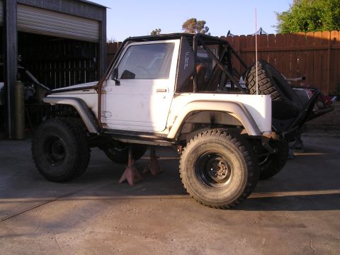 1987 Suzuki Samurai - This modified Suzuki Samurai is parked outside a garage with a roll cage and large off-road tires.
