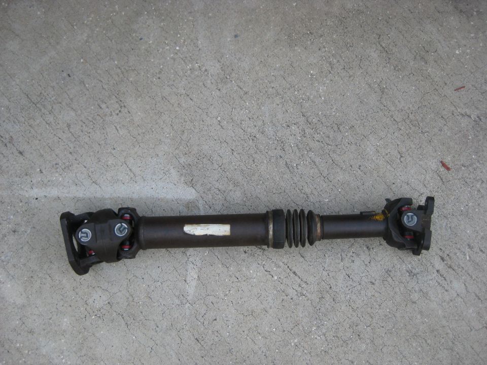 Driveshaft with U-joints and a rubber boot, lying on concrete. White label on shaft