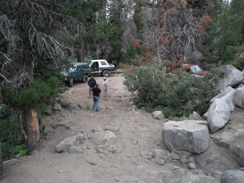 1991 Ford Bronco - Several off-road vehicles are parked on a rocky trail surrounded by dense forest.