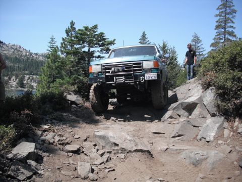 1991 Ford Bronco - A teal Ford Bronco navigates a challenging rocky trail with a person standing on the side.