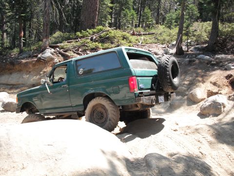 1991 Ford Bronco - A green Ford Bronco navigates a challenging off-road path with large rocks and trees.